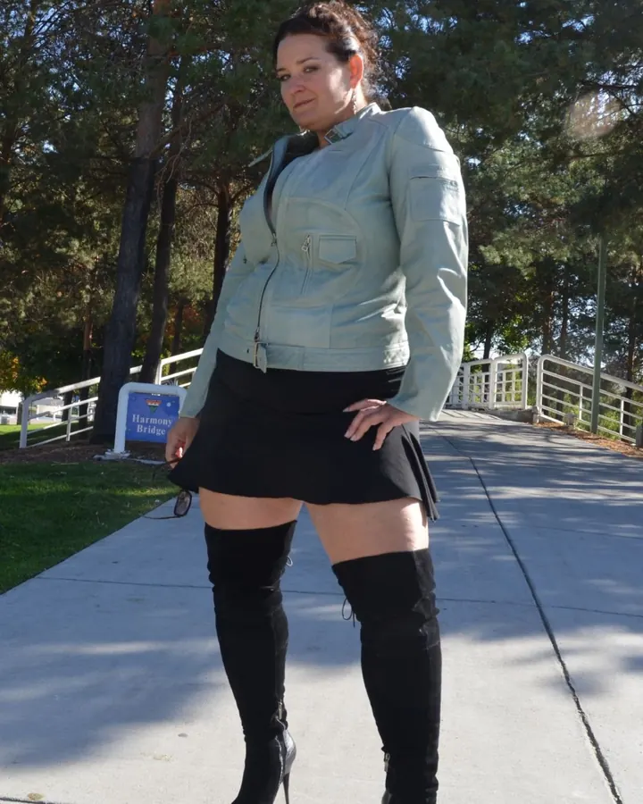 Femme en veste en cuir clair, jupe noire et bottes hautes, posant en extérieur près d’un pont.