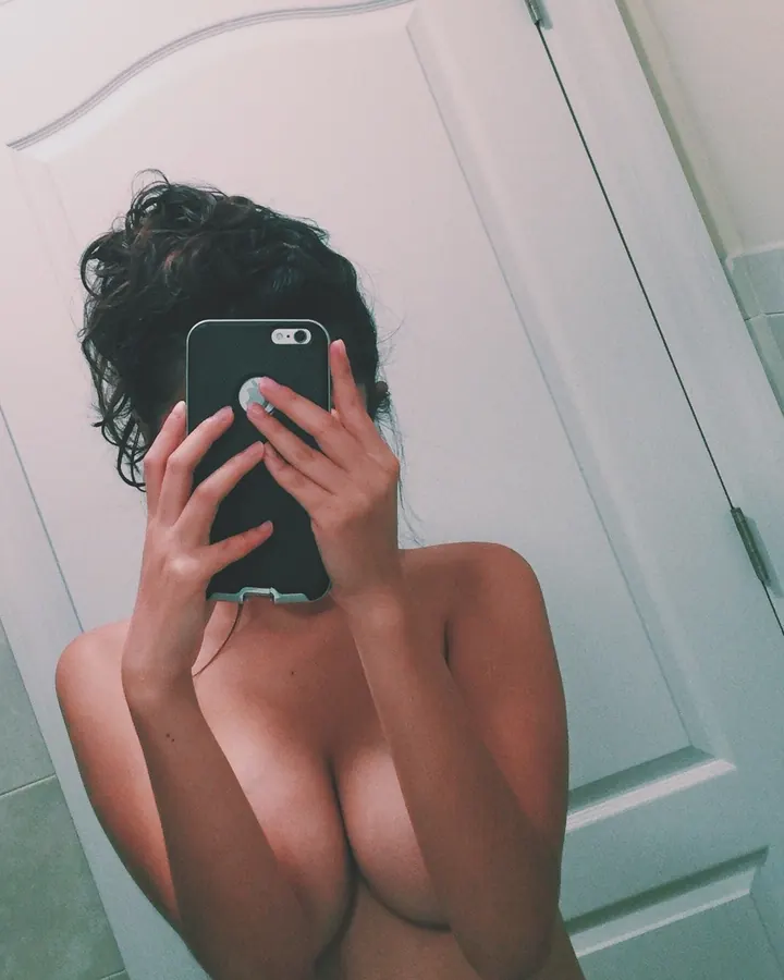 Femme nue prenant un selfie dans une salle de bain, le téléphone masquant son visage.