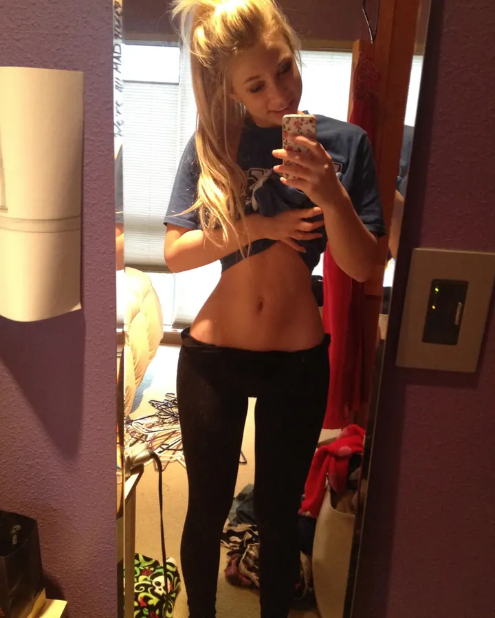 Femme blonde, en haut bleu et leggings noirs, prenant un selfie devant un miroir dans une chambre.