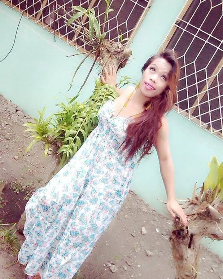 Femme arabe de 35 à 45 ans en robe blanche fleurie, posant dehors près d’un mur turquoise et de plantes.