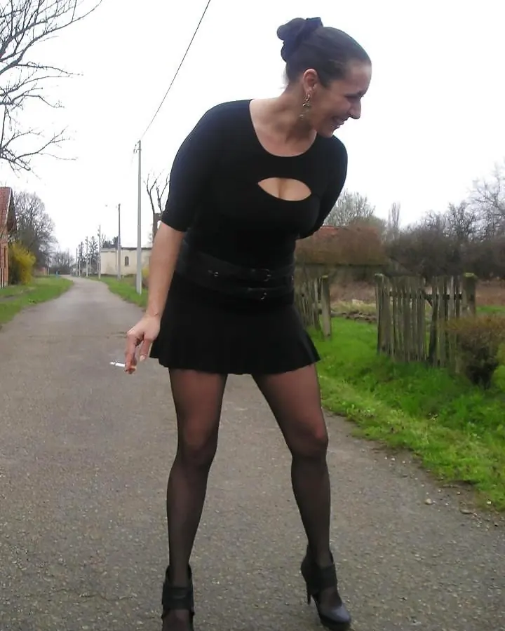 Femme arabe de 45 à 60 ans, en tenue noire, debout sur un chemin rural, tenant une cigarette.